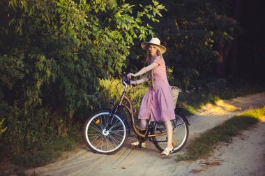 Eğleniyor bir parkta güzel bir sepet çiçekli tutan bir bisiklet ile güzel bir pembe elbise giyen güzel kız. Vintage manzaralar. Retro görünüm, Bisiklet ve sepet çiçekli güzel sarışın