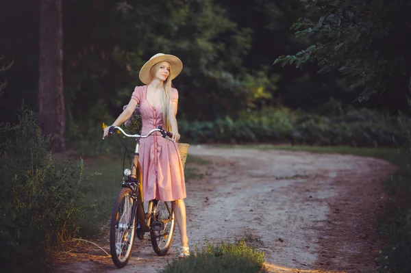 Όμορφη κοπέλα φορώντας ένα ωραίο ροζ φόρεμα διασκέδαση σε ένα πάρκο με ένα ποδήλατο που κρατά ένα όμορφο καλάθι με λουλούδια. Vintage τοπία. Όμορφη ξανθιά με ρετρό εμφάνιση, ποδήλατο και καλάθι με λουλούδια — Φωτογραφία Αρχείου