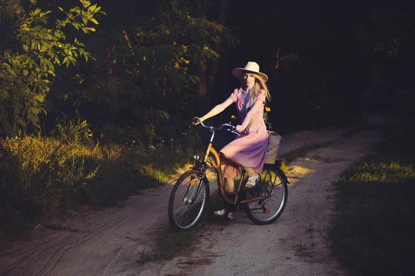 Hermosa chica con un bonito vestido rosa divirtiéndose en un parque con una bicicleta sosteniendo una hermosa canasta con flores. Paisajes antiguos. Bonita rubia con look retro, bicicleta y cesta con flores — Foto de Stock
