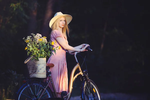 Hermosa chica con un bonito vestido rosa divirtiéndose en un parque con una bicicleta sosteniendo una hermosa canasta con flores. Paisajes antiguos. Bonita rubia con look retro, bicicleta y cesta con flores —  Fotos de Stock