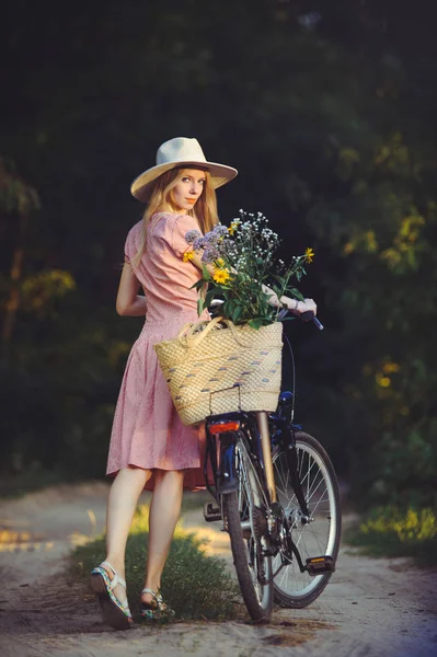 Hermosa chica con un bonito vestido rosa divirtiéndose en un parque con una bicicleta sosteniendo una hermosa canasta con flores. Paisajes antiguos. Bonita rubia con look retro, bicicleta y cesta con flores —  Fotos de Stock