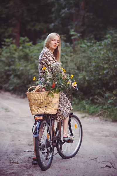 Hermosa chica con un bonito vestido divirtiéndose en un parque con una bicicleta sosteniendo una hermosa canasta con flores. Paisajes antiguos. Bonita rubia con look retro, bicicleta y cesta con flores — Foto de Stock