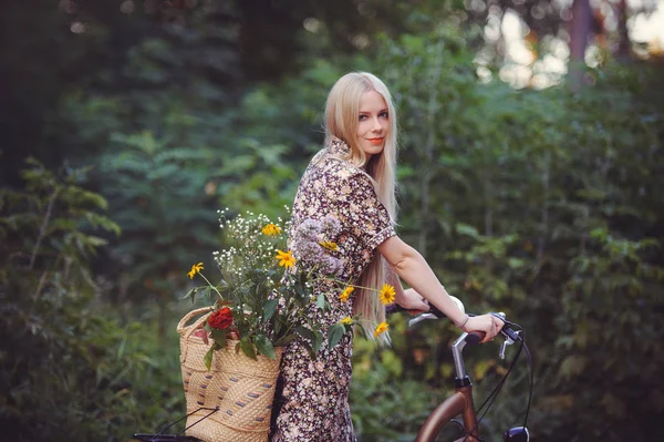 Hermosa chica con un bonito vestido divirtiéndose en un parque con una bicicleta sosteniendo una hermosa canasta con flores. Paisajes antiguos. Bonita rubia con look retro, bicicleta y cesta con flores — Foto de Stock