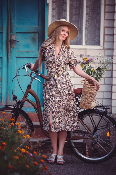 Hermosa chica con un bonito vestido que se divierte en una casa de campo con una bicicleta, sosteniendo una hermosa canasta con flores. Paisajes antiguos. Bonita rubia con un look retro — Foto de Stock