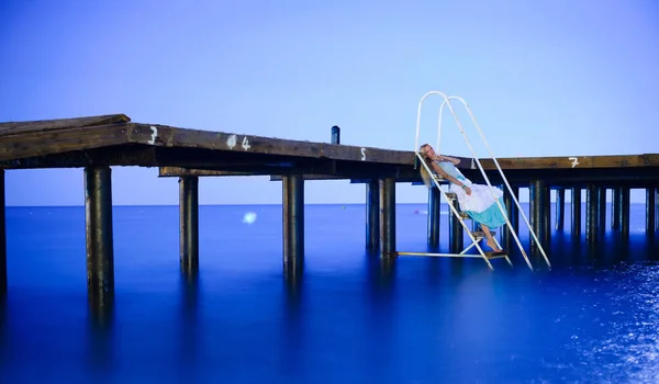 Uma mulher solitária e pensativa sentada nos degraus do cais e admirando a noite azul. Mulher bem vestida em um vestido azul branco, recreação e turismo, área de resort — Fotografia de Stock