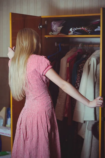 Blonde woman looks in a closet, dressed in a red dress, stands in an old house, a simple home image — Stock Photo, Image