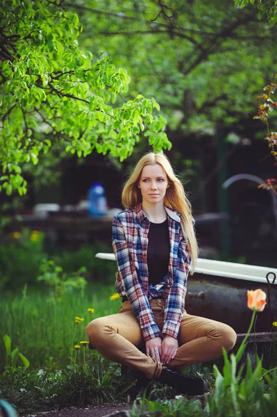 Niedliche blonde Frau sitzt in einem heimischen Garten, auf einem Hintergrund von vielen grünen Bäumen und Tulpen, ein schönes Porträt eines rustikalen Stils, ein einfaches Hausbild — Stockfoto