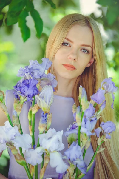 Attractive young girl with blonde dren hair and natural make-up smelling blue purple iris flowers on a background outdoors, tenderness and softness on a nature background — Stock Photo, Image