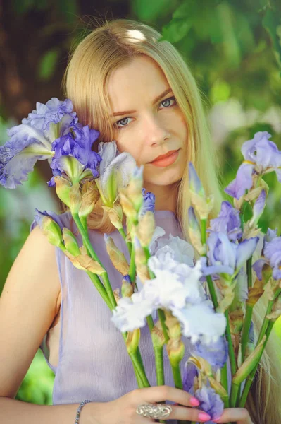 Muchacha joven atractiva con cabello rubio y maquillaje natural oliendo flores de iris azul púrpura sobre un fondo al aire libre, ternura y suavidad sobre un fondo natural —  Fotos de Stock