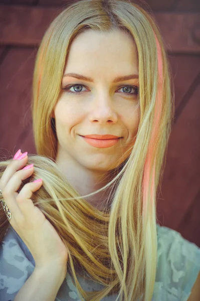 Atrakcyjne skromna młoda blond kobieta grając z włosów na czerwonym tle drewniane malowane jej włosy w różowe paski paski, w niebieskiej sukience nieśmiały i ciekawy — Zdjęcie stockowe
