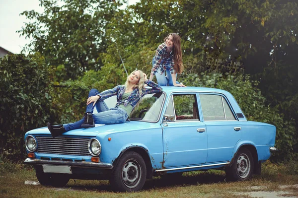 Весела Приваблива дві молода блондинка і брюнетка позує на капоті старого іржавого автомобіля, одягненого в джинси і сорочки на природі — стокове фото