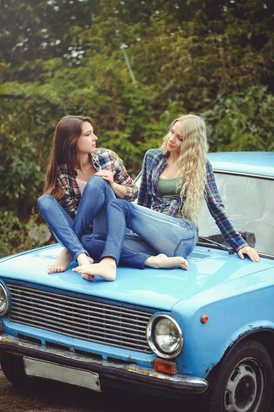 Cheerful Attractive two young blonde girl and brunette posing on the hood of an old rusty car, dressed in jeans and shirts on a nature background — Stock Photo, Image