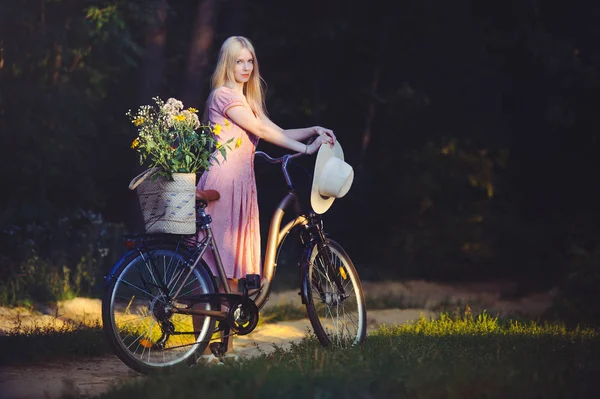 Hermosa chica con un bonito vestido rosa divirtiéndose en un parque con una bicicleta sosteniendo una hermosa canasta con flores. Paisajes antiguos. Bonita rubia con look retro, bicicleta y cesta con flores —  Fotos de Stock