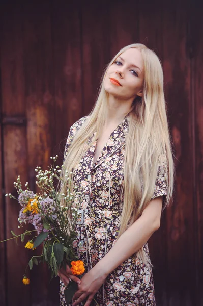 Hermosa mujer joven en vestido de verano con puntos sobre fondo rojo de madera vieja, mirando a la cámara. estilo rústico, bonito con flores silvestres en las manos — Foto de Stock