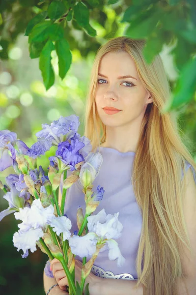 有吸引力的年轻女孩与金发和自然化妆嗅到的蓝色紫色虹膜花在户外背景下, 柔软和柔软的自然背景 — 图库照片