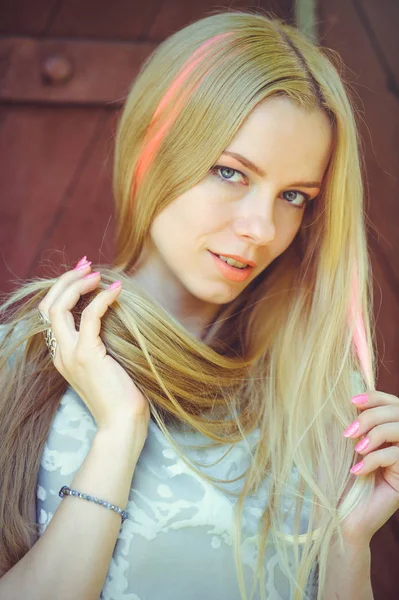 Atraente modesto jovem loira brincando com o cabelo no fundo de madeira vermelho seu cabelo pintado em listrado listrado rosa, em vestido azul tímido e curioso — Fotografia de Stock