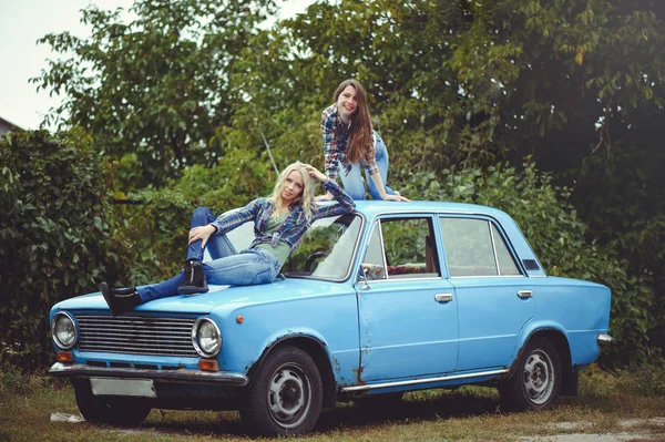 开朗迷人的两个年轻的金发女孩和黑发摆在旧生锈的汽车的敞篷, 穿着牛仔裤和衬衫的性质背景 — 图库照片#