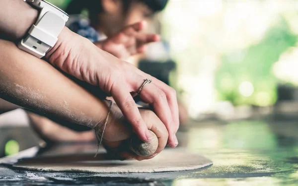 Adult hand is teaching a child to make pizza. A mother is teaching her son to roll the bread on kitchen table. A Family is cooking homemade pizza.