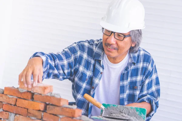 Old construction worker is laying down brick wall with cement, w