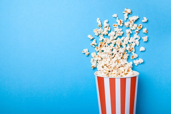 Palomitas de maíz derramadas y cubo de papel en tira roja sobre fondo azul. Copiar espacio para texto —  Fotos de Stock