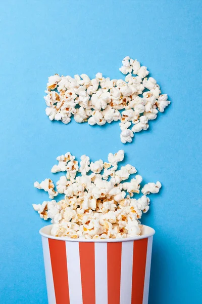 Verschüttetes Popcorn in Form eines Pfeils nach rechts und Papiereimer in rotem Streifen auf blauem Hintergrund. — Stockfoto