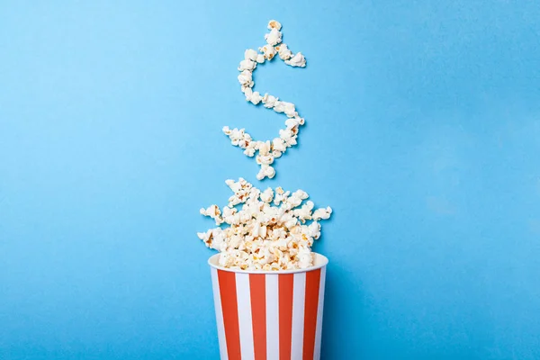 Konzept Eintrittspreise oder Kinokasse. verschüttetes Popcorn in Form eines Dollarzeichens aus einem Papiereimer in einem roten Streifen auf blauem Hintergrund. — Stockfoto