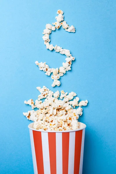 Konzept Eintrittspreise oder Kinokasse. verschüttetes Popcorn in Form eines Dollarzeichens aus einem Papiereimer in einem roten Streifen auf blauem Hintergrund. — Stockfoto