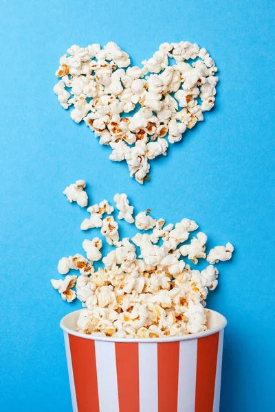 Ich schaue gerne Filme. verschüttetes Popcorn in Herzform und Papiereimer in einem roten Streifen auf blauem Hintergrund. — Stockfoto