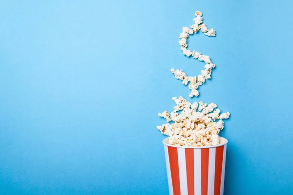 Konzept Eintrittspreise oder Kinokasse. verschüttete Popcorn in Form eines Dollarzeichens aus einem Papiereimer in einem roten Streifen auf blauem Grund. Kopierraum für Text — Stockfoto