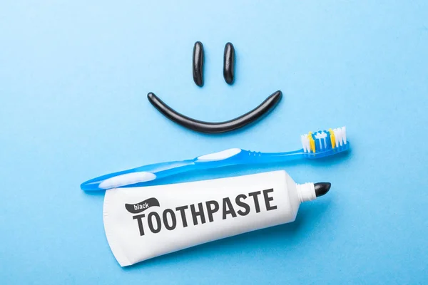 Black toothpaste from charcoal for white teeth. Toothpaste in the form of  smile on the face,  tube and  toothbrush on  blue background. — Stock Photo, Image