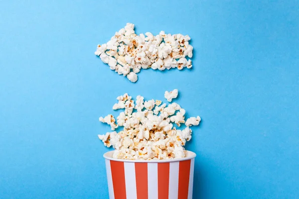 Palomitas de maíz derramadas en forma de flecha a la izquierda y cubo de papel en tira roja sobre fondo azul . — Foto de Stock