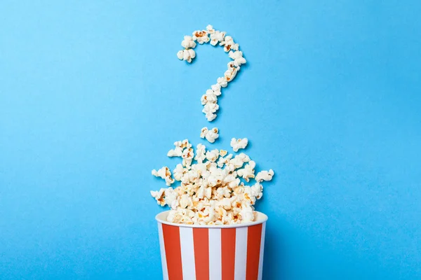 Concepto de qué película elegir. Palomitas de maíz derramadas en forma de signo de interrogación y un cubo de papel en tira roja sobre fondo azul . — Foto de Stock