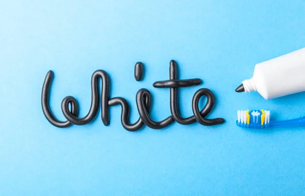 Черная зубная паста из угля для белых зубов. Слово белое от черной зубной пасты, тюбик и зубная щетка на синем фоне . — стоковое фото
