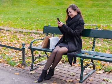 Güzel bir kız bankta oturuyor, telefonuyla sosyal ağlara bir mesaj yazıyor, güneşli bir sonbahar günü parkta, alışveriş sonrası dinlenen bir kadın..