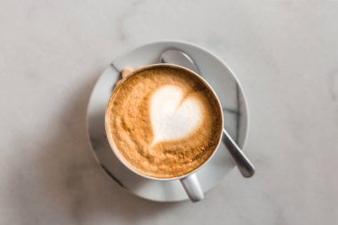 Bir fincan kapuçino, beyaz mermer masa üzerinde latte sanatı, beyaz seramik bardak, üst manzara. Kafe kültürü.