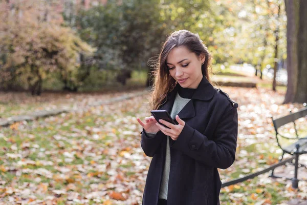 Mujer morena con su teléfono escribe un mensaje en las redes sociales, día de otoño soleado en el parque, relajante, después de la mujer de negocios de trabajo — Foto de Stock