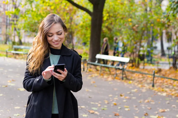 Mujer morena con su teléfono escribe un mensaje en las redes sociales, día de otoño soleado en el parque, relajante, después de la mujer de negocios de trabajo — Foto de Stock