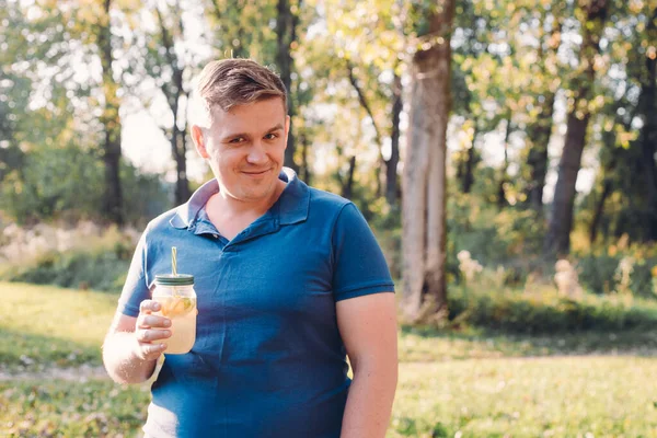 Man in casual wear posing over green city park on sunny warm day, holding cup of lemonade and looking to camera with smile. Vitamins and regular life concept