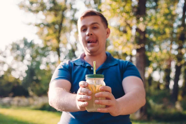 Man in casual wear posing over green city park on sunny warm day, holding cup of lemonade, licking lips and looking to camera. Vitamins and regular life concept