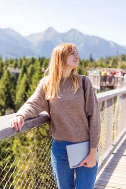 Uçurumun kenarında duran tabletli genç bir kadın. Seyahat ve turizm konsepti. Copyspace Dağların arka planında duran kızıl saçlı genç bir kadın