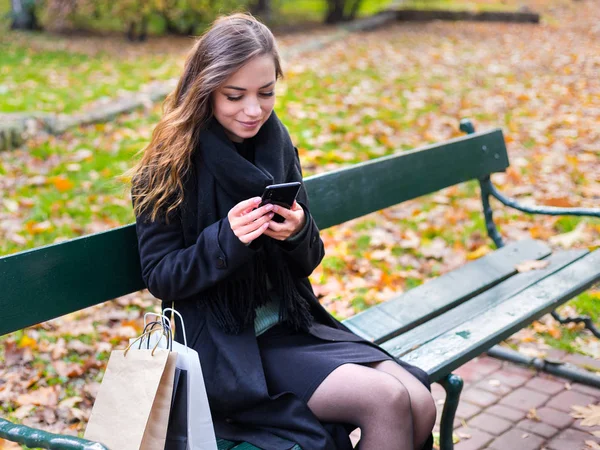 Hermosa chica sentada en un banco, mujer con su teléfono escribe un mensaje en las redes sociales, día de otoño soleado en el parque, mujer relajante después de ir de compras — Foto de Stock