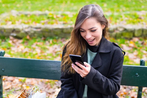 Hermosa chica sentada en un banco, mujer morena con su teléfono escribe un mensaje en las redes sociales, día soleado en el parque, relajante, mujer de negocios después del trabajo — Foto de Stock