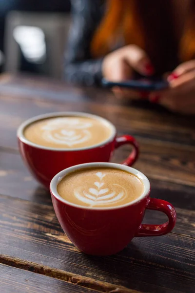 Twee rode kopjes koffie op houten ondergrond met latte art. Tafel in café. Concept van gemakkelijk ontbijt. Kleine en grote keramische bekers. Bovenaanzicht. Meisje met telefoon op de achtergrond — Stockfoto