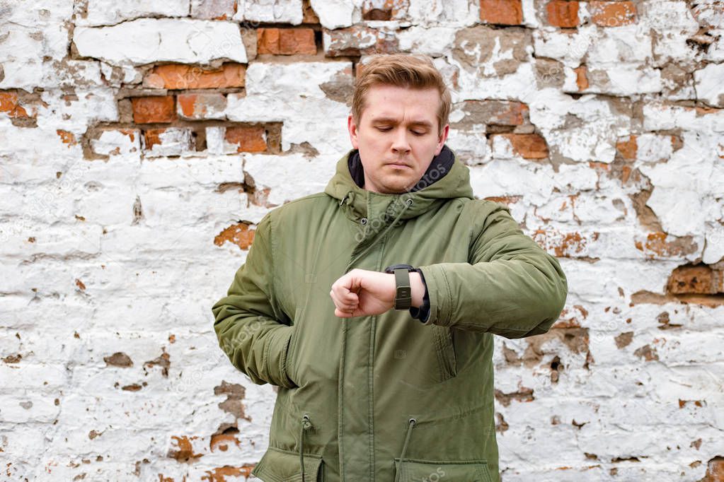 Man checking the time on his wrist watch. Handsome young man stay against white and red old brick wall, winter time