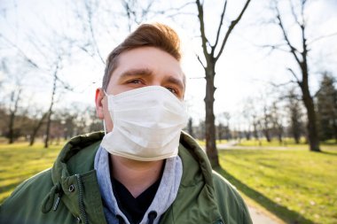 Şehir parkının arka planında tıbbi maske takan bir adamın portresi. Corona virüsü salgını. Hava kirliliği, zatürree salgını, sis veya salgın hastalık kavramı