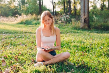 Beyaz tişörtlü, sarı saçlı genç kız gün batımında tarlada kitap okuyor..