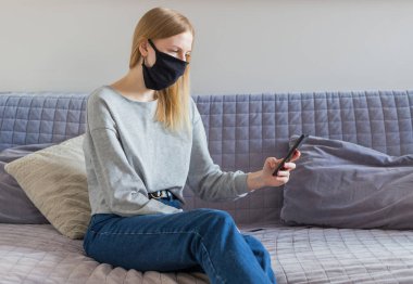 Coronavirus. Kadın koruyucu maske taktığı için karantinada, evden çalışıyor ve koltukta cep telefonu kullanıyor. Salgın sağlık konsepti