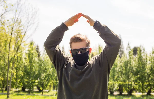 Человек в защитной черной маске делает домашний знак в городском парке. Принято. Символ дома, выполненный руками на природном фоне. Концепция строительства Гая удерживает крышу над головой — стоковое фото