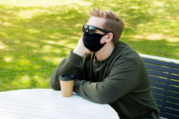 Tristeza Hombre con una máscara negra protectora sentado en una mesa en un café. Protección contra virus. Hombres con máscara tomando café en un parque de la ciudad. Copiar espacio . — Foto de Stock