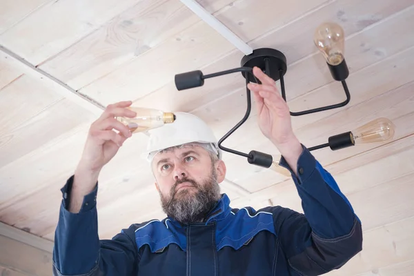 a male electrician performs in a white hard hat and with a beard performs the replacement of a light bulb in the ceiling chandelier. repair of ceiling lamp